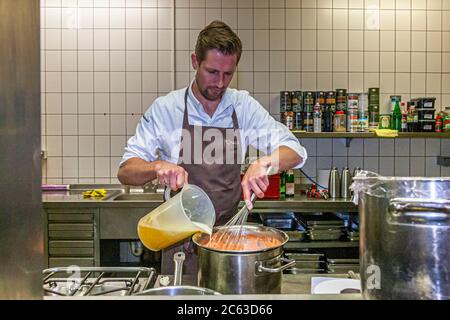 German two-star chef Michael Kempf stirs a pot with focused attention on the gas stove of the Parkhotel Ahrensburg kitchen, Germany. More pots and pans stand within reach on the stainless-steel counter around him. Kempf is head chef of Berlin's two-Michelin-star restaurant Facil at Hotel Mandala. Stock Photo
