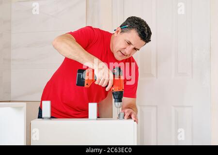 middle aged white man screwed the cabinet legs into wooden furniture with cordless screwdriver closeup. assembling wooden kitchen for the house Stock Photo