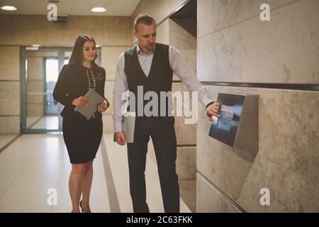Two business colleagues near the lifts room indoors of a contemporary office skyscraper: man entrepreneur with a laptop is using the touch screen Stock Photo