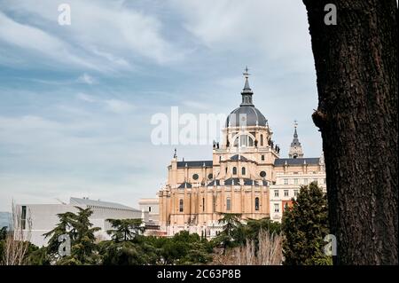 Exterior architecture of marvellous Almudena Cathedral seen from behind trees against cloudy sky in Madrid in Spain Stock Photo