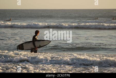 A young male surfer walks into the ocean at sunset Stock Photo