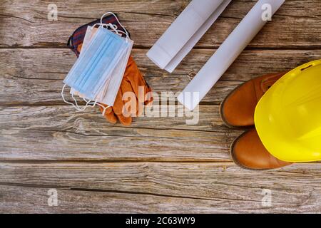 Industry worker protective and surgical mask, workwear safety boots leather gloves yellow hard hat on architects working with blueprints in the office coronavirus pandemic covid-19, Stock Photo