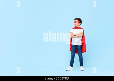 Superhero boy in red mask and cape with arms crossed thinking and looking at copy space aside isolated light blue background Stock Photo