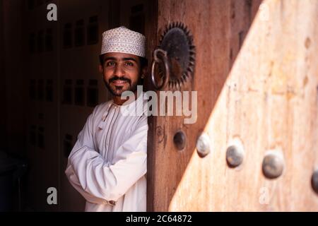 Nizwa / Oman - February 15, 2020: Portrait of young handsome Omani man smiling next to wooden door in Nizwa Souq Stock Photo