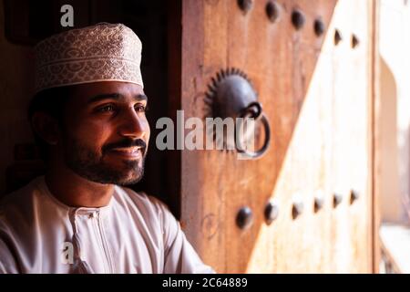 Nizwa / Oman - February 15, 2020: Portrait of young handsome Omani man smiling next to wooden door in Nizwa Souq Stock Photo