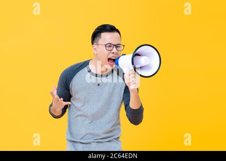 Angry young Asian man in gray top and eyeglasses shouting into megaphone isolated on yellow background Stock Photo
