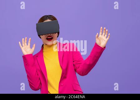 Shocked woman in colorful attire watching 3D simulation video from virtual reality or VR glasses on purple background Stock Photo