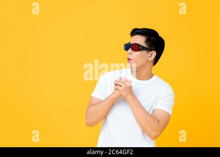 Excited young handsome Asian man wearing 3D movie glasses looking at empty space with hands together isolated on yellow background Stock Photo