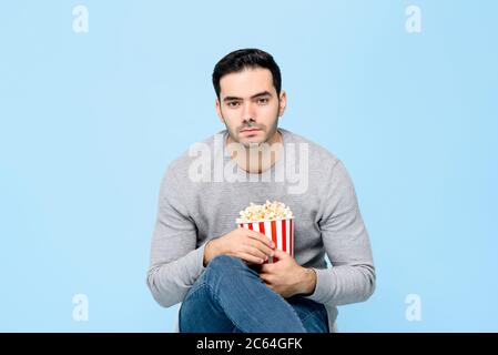 Bored young man holding popcorn sitting and watching movie isolated on light blue background Stock Photo