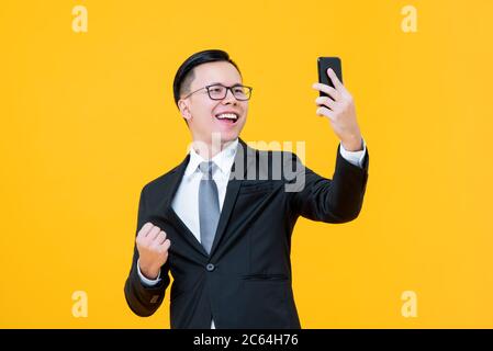 Portrait of happy confident Asian businessman taking a selfie doing a success closed fist gesture in isolated studio yellow background Stock Photo