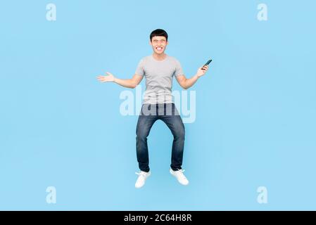 Fun full length portrait of happy smiling Asian man jumping in mid-air while holding smartphone in isolated studio blue background Stock Photo