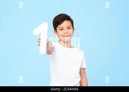 Portrait of smiling young Asian boy holding and showing number one cutout in isolated studio blue background for education concept Stock Photo