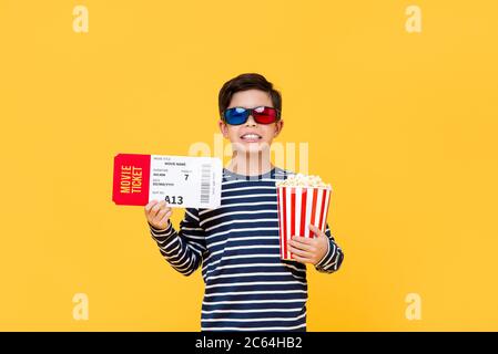 Fun portrait of happy young Asian boy wearing 3D glasses holding popcorn and movie ticket in isolated studio yellow background