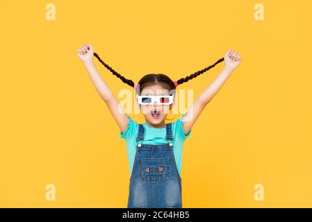 Fun Portrait of excited young girl wearing 3D cinema glasses with both arms raised pulling her hair in isolated studio yellow background