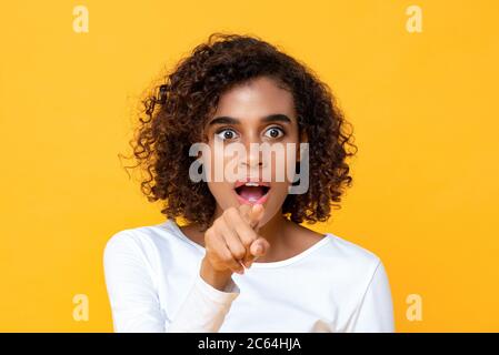 Close up portrait of attractive young African American woman looking shocked while pointing finger at front in isolated studio yellow background Stock Photo