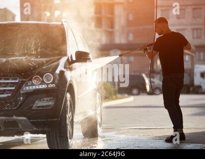 Silhouette of a strong man holding a hose and washing his black car with a water jet, water drops and side of the car are shining on the setting sun Stock Photo