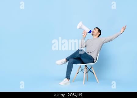 Portrait of young handsome Caucasian man sitting and cheerfully announcing on megaphone with one arm raised in isolated studio blue background Stock Photo