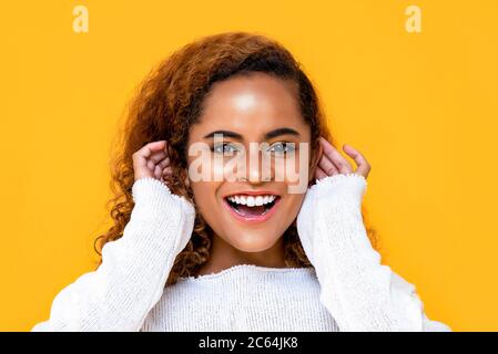Close up portrait of cheerful young African American woman smiling while touching her ears with both hands in isolated studio yellow background Stock Photo