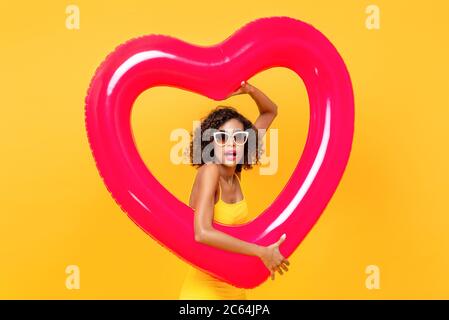 Portrait of surprised African American woman wearing sunglasses holding heart shape balloon in isolated studio yellow background Stock Photo
