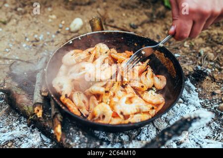 The cook stirs with a fork the sizzling shrimps that are fried in a pan in oil - seafood in a spicy garlic sauce