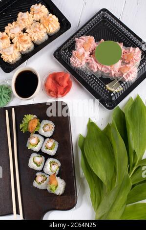 Sushi rolls set served on dark wooden board and plastic boxes. Restaurant food delivery in take away boxes. Gray background with green leafs