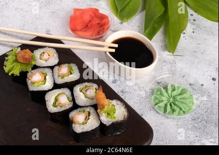Rolls  with fried shrimp and salad. Sushi rolls set served on dark wooden board with wasabi, soy sauce and ginger. Gray background with green leafs