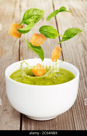 fresh leaves and croutons falling into green soup bowl on wooden table Stock Photo