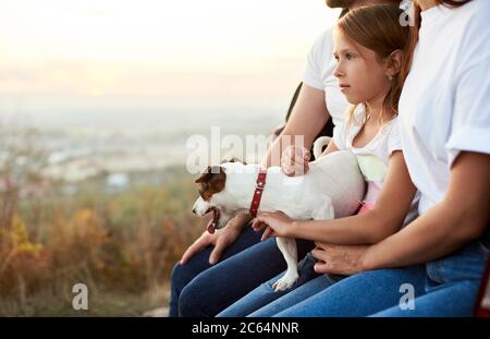Close-up shot of the girl with her pet dog on her laps, sitting in the car trunk between her parents, family wearing white t-shirt and jeans enjoys a beautiful view from the hill Stock Photo