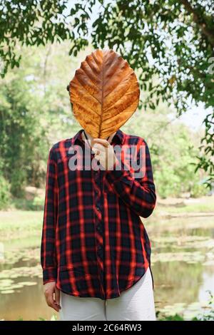 Young woman in a checkered shirt in black and red covers her face with a large, dried leaf of a tree. Stock Photo