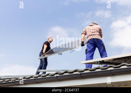 Happy electrical engineers mans are holding solar panel on roof against blue sky. Development sun alternative energy technology. Ecological concept. Stock Photo