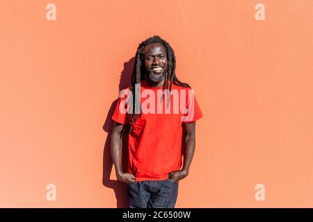 Portrait of Black man with dreadlocks standing in front of orange wall, smiling at camera. Stock Photo