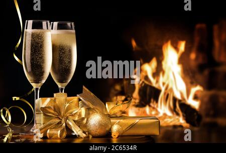 Celebration toast with champagne. Two champagne flutes with gold gift boxes and streamers in front of fireplace Stock Photo