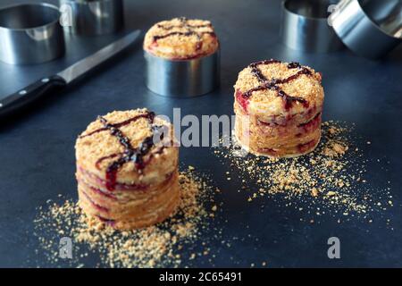 Round portions of homemade honey cake with sour berry layers on dark table. Molding rings on a background. Stock Photo
