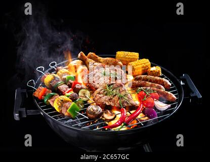 Assorted delicious grilled meat with vegetables on barbecue grill with smoke and flames isolated on black background Stock Photo