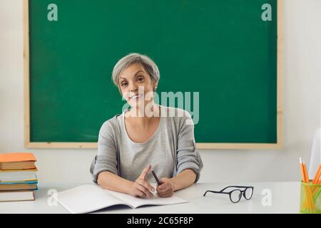 The teacher is looking at the camera online. Female senior teacher teaches students remotely using computer video chat in classroom. Stock Photo