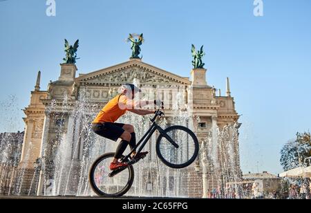 Extreme sportsman performing dangerous ride on mountain bike back wheel in front of fountain and opera house, side view, horizontal snapshot Stock Photo