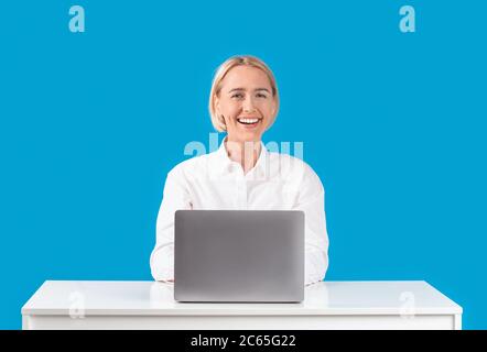 Cheerful mature business lady working with laptop at desk on blue background Stock Photo