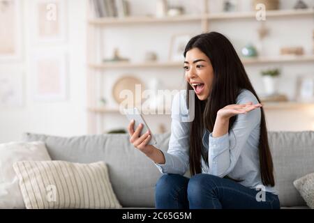 Unexpected Message. Asian girl holding smartphone at home, emotionally reacting to news Stock Photo