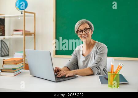Teacher school university online. Woman senior teacher speaks with students has web chat using laptop computer sitting at desk in classroom. Stock Photo