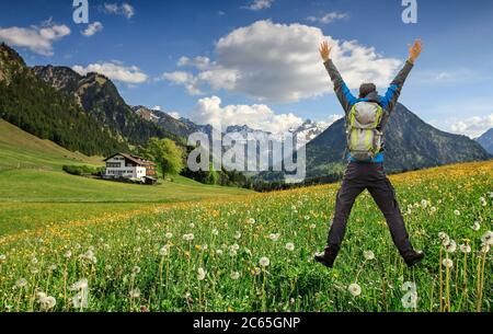 Happy succesful Hiker with Backpack jumping with raised arms on Hawkbit Flower meadow. Snow covered mountains and traditional house. Bavaria, Alps Stock Photo