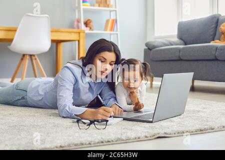 Work online. Woman working from home using laptop computer lying on livingroom carpet with curious daughter. Stock Photo
