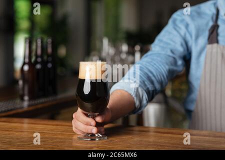 Serving client with drinks. Barman in apron gives glass of dark beer with foam on wooden bar Stock Photo
