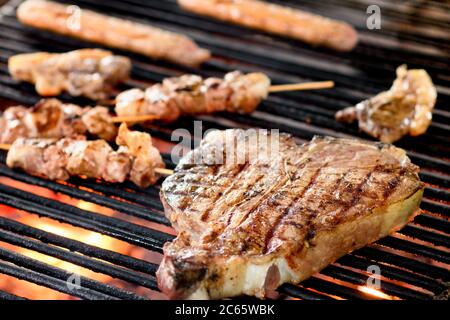 Grilling a beef Florentine steak on a barbecue fire with kebabs and sausages in close up with focus to the steak over the hot coals Stock Photo