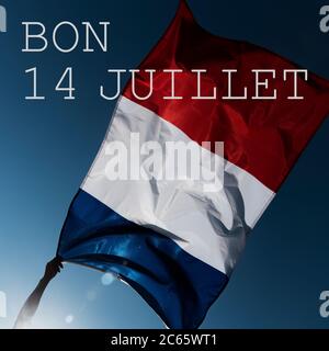 man holding a French flag waving on the wind and the text bon 14 juillet, happy 14 july, the national day of France written in French, against the blu Stock Photo