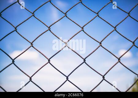 Blue sky with clouds through rusty wire mesh fence. Protection border and forbidden line. Blur background, texture. Close up view of link cage, wallpa Stock Photo