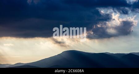 Cloudscape at sunset. Heavy dark clouds on sky over mountains background. Sunbeams through fluffy clouds color the blue sky and the hills black silhou Stock Photo