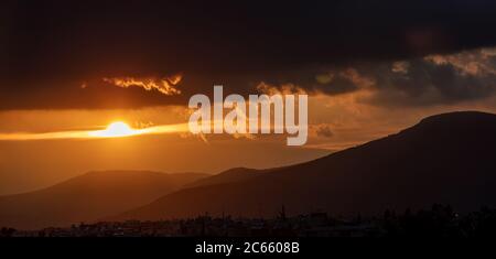 Sunset, sunrise through and behind the clouds over the mountains background. Sunbeams color the sky the village houses and the hills black silhouette. Stock Photo