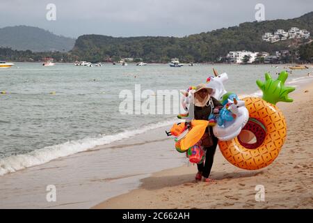 Female beach seller walking on the sands with range of colourful inflatables, including rubber rings, for sale at Koh Samui, Thailand Stock Photo