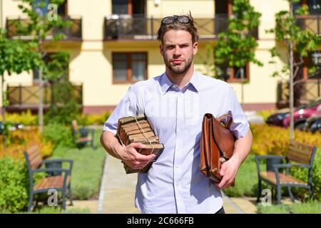 Man with briefcase and pile of old books in yard on sunny day. Academic style and studying concept.Student or young businessman with glasses and strict face outdoor. Stock Photo