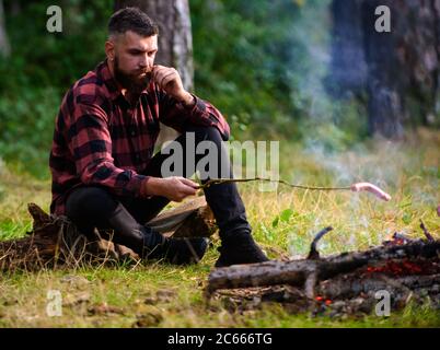 Guy with tired face and lonely at picnic or barbecue. Man, hipster, hiker roasting sausages on stick on bonfire in forest. Hungry wanderer concept. Hipster with beard cooking food. Stock Photo